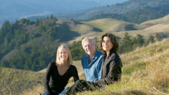 Three people sit on a grassy hillside overlooking the scenic landscape of rolling hills and forests. The people, two women and one man, are relaxed and smiling, enjoying the natural surroundings under a clear sky with a stunning view of Mount Tam in the distance.