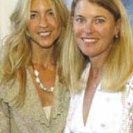 Two smiling women with long, blonde hair posing together. The woman on the left is wearing a patterned blouse and a chunky necklace, while the woman on the right is dressed in a white top with lace detail. Both are standing indoors in what seems like a cozy cabana with a blurred background.