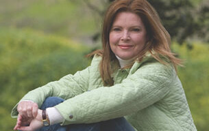 A woman with long brown hair sits outdoors in a green field, wearing a light green quilted jacket and blue jeans. She smiles softly, leaning on her bent knee with her arms crossed casually. In the serene background of blurred greenery, Leigh Glaser's presence adds a subtle elegance to the scene.