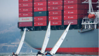 Three small sailboats navigate near a large container ship labeled "K Line," stacked high with red cargo containers. The scene, reminiscent of old Knarr voyages, is set in a body of water amidst a foggy backdrop, with the coastline visible in the distance.