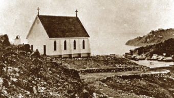 A sepia-toned photograph shows a small, one-room church with two crosses on its roof, situated on a rocky hillside overlooking a body of water. The background features distant hills and a few buildings near the water's edge.