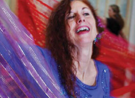 A woman smiles brightly while dancing, surrounded by flowing, colorful fabric. She wears a blue top and has wavy, reddish-brown hair. The background features soft, artistic scenery reminiscent of Middle Eastern landscapes, and the word "PLAY" is visible at the top left.