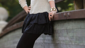 A person leans casually on a wooden railing outdoors in Kentfield, wearing a light-colored long-sleeve top, a black tiered skirt, black leggings, and black heeled shoes. They have short, dark hair with a headband and a wide bracelet on one wrist. Lush green foliage is in the background, reminiscent of Ghazaleh Khalifeh’s serene landscapes.
