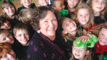A smiling woman with curly hair in the center is surrounded by a group of happy children, who also appear to be smiling. The children are dressed in festive, colorful clothing with some wearing green wigs or accessories. The cheerful and lively environment radiates the essence of making a difference.