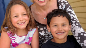 A woman with short blonde hair and a black and white floral top is sitting outside, smiling with two children. The girl on her left is wearing a white dress with pink flowers, while the boy on her right, in a black T-shirt with a white logo, looks like he just had an attitude adjustment.