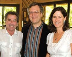 Three people are smiling and standing close together in front of large windows at the McEvoy Ranch Harvest Party. The person on the left is wearing a white shirt, the person in the center is wearing glasses and a striped shirt, and the person on the right is wearing a white blouse.