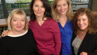 Four women are standing together inside a room with large windows in Mill Valley. They are posed closely and smiling towards the camera. The woman on the left has short blonde hair, the second woman has dark brown hair, the third has long blonde hair, and the fourth has curly hair.