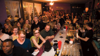 A crowded bar with patrons standing and sitting along a long counter. Many people, including book lovers, are smiling and socializing. A bartender on the right is preparing drinks. The bar is well-lit and decorated with various literary-themed items and bottles.