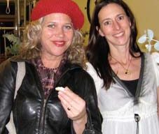 Two women are standing indoors, smiling at the camera. The woman on the left embodies a chic style with her red beret, black leather jacket, and plaid shirt, holding a small item. The woman on the right is wearing a white blouse with a black undershirt and a pendant necklace.