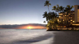 Sunset beach scene with gentle waves washing ashore, silhouettes of palm trees, and a well-lit resort building on the right. The sky transitions from deep blue to the warm hues of the setting Sun, casting a serene glow over the water.