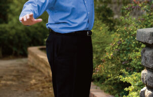 A person wearing a blue shirt and black pants is posing outdoors in Mill Valley, with one leg slightly raised and arms extended, appearing to balance or dance. They are smiling and standing on a brick path with greenery and a building in the background, reminiscent of an Alan Scofield performance.