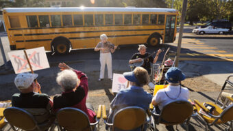 A group of seated elderly people at a roadside bus stop watch two musicians performing. One musician plays a violin and the other a banjo, their melodies adding rhythm to life. A bright yellow school bus is visible in the background. Some audience members hold signs, one of which is crossed out, framed by towering Redwoods nearby.
