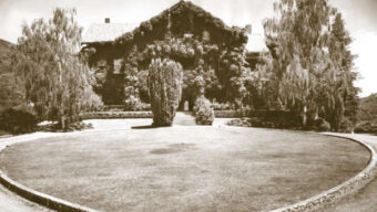 A sepia-toned image of a large, ivy-covered house with a stone pathway leading to the entrance. This enchanting abode, known as the Ralston L. White Retreat, is flanked by tall trees and surrounded by a landscaped lawn and circular driveway. Hills loom in the background under a partly cloudy sky.