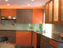 Modern kitchen with confident orange and gray cabinets, stainless steel appliances, and a dark countertop. The kitchen features a built-in gas stove, overhead lights, and a small, round dining table with two chairs. A fruit bowl is positioned near the sink.