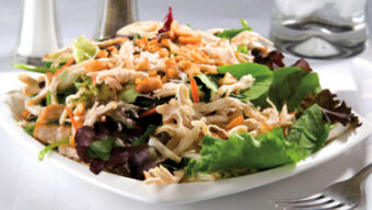A fresh Chinese Chicken Salad on a white plate featuring shredded chicken, mixed greens, and nuts. In the background, there is a salt and pepper shaker, a glass of water with ice, and a fork placed on the right side of the plate on a white surface.
