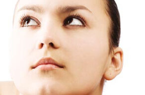 A young person with fair skin and dark brown, slicked-back hair is gazing upward thoughtfully. Resting their chin on their hands against a plain white background, they exude natural beauty. With light makeup and long eyelashes, they look serene and contemplative, reminiscent of the tranquil landscapes of Marin County.