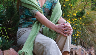 Lisa Kaiser, a woman with long hair and a green scarf draped over her shoulders, sits on a stone ledge in a garden. She is wearing a patterned top and light-colored pants, smiling at the camera with her hands resting on her knees. Greenery and flowers surround her.