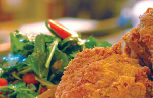 A close-up of a plate with crispy fried chicken, the epitome of comfort food, served next to a fresh salad with mixed greens and sliced red bell peppers. The background is softly blurred, drawing focus to the appetizing meal. Ideal for a cozy evening in Mill Valley.