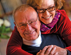 An elderly couple, Jim and Nancy Lange, both wearing glasses, share a joyful moment indoors. Nancy Lange, in a red sweater and scarf, hugs Jim from behind as he wears a dark sweater with a white undershirt. They both smile warmly at the camera.