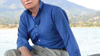 A man with white hair and a moustache, dressed in a blue shirt and beige pants, sits on a wooden surface outdoors with a lake and mountains in the background. Doug Ferguson appears relaxed, with one arm resting on his knee and the other on the wood. This peaceful citizen enjoys nature's serenity.