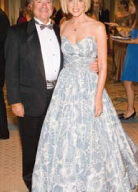 A man in a black tuxedo and bow tie stands beside a woman in a strapless, floor-length, light blue and white gown. They are smiling and radiate love as they pose for the camera at a formal event, with other guests in the background.