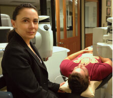 A woman in a black blazer sits next to a person lying on a medical examination bed, receiving an incredible eye exam. The room contains ophthalmic examination equipment, and the individual lying down is wearing a red shirt and resting their head on a cushion.
