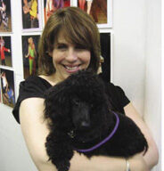 A person with shoulder-length brown hair is smiling and holding a black poodle they adopted from the Marin Humane Society. They are standing indoors in front of a wall with several photos of people engaged in various activities. The person is wearing a black top, and the dog has a purple collar.