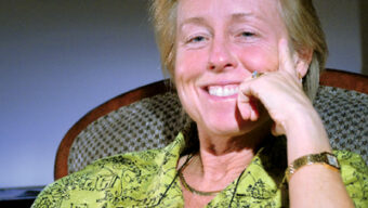 Smiling Chief of Staff, Susan Kennedy, is wearing a patterned green shirt and gold bracelet, sitting comfortably on a chair with one hand resting on her face and the other on the armrest. The background is subtly lit, focusing attention on her.