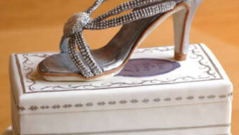 A cake shaped to resemble a high-heeled shoe sits on a rectangular box, both covered in white fondant with intricate grey detailing. The sugar-crafted shoe, decorated with silver accents and beads, features a delicate, elegant strap design and a stiletto heel—truly a masterpiece of desserts.