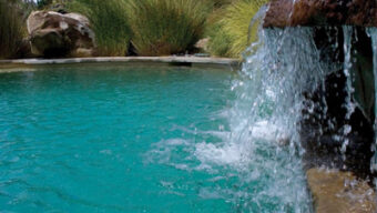 A serene pool with turquoise water featuring a small, cascading waterfall flowing over natural rock steps. Surrounding the pool are large rocks and lush greenery providing a tranquil and inviting atmosphere, showcasing one of the latest pool trends in outdoor design.