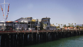 A sunlit pier with wooden supports extends over calm water in Santa Barbara. The pier is lined with buildings, including a white two-story structure with a patio where you can savor delightful Santa Barbara dining. Colorful flags flutter on a pole. In the background, there are palm trees and distant mountains under a clear sky.