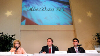 Three people are seated at a table covered with a blue tablecloth, participating in a panel discussion under a large screen displaying "Election 08." The panelists have microphones and nameplates, with star decorations on the wall behind them, signaling the intense State Election atmosphere.