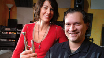 A woman holding a trumpet and a man smiling pose together in a room filled with audio equipment and speakers. The woman, wearing a red top, exudes Film Magic while the man, dressed in a dark shirt, radiates charm.