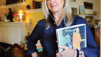 A woman with long blonde hair stands in a warmly-lit, cozy room. She is wearing a navy blue shirt and white pants, accessorized with a large belt and silver jewelry. The woman is holding a book about family history, the cover featuring a person in an orange outfit.