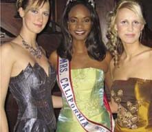 Three women in elegant dresses stand together, smiling. The woman in the center wears a tiara and sash that reads "Mrs. California." Each dress is uniquely stylish with intricate designs. They appear to be at a formal event or pageant, possibly supporting the Make-A-Wish Foundation.