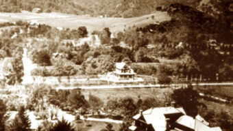 A sepia-toned historical photograph depicts two large houses surrounded by trees and greenery, set within a mountainous landscape. The houses, reminiscent of early Marin architecture, are separated by a road, and the hills in the background add depth to this garden-like scene.