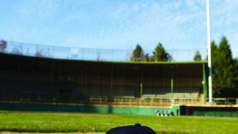 A baseball and a navy blue cap with the word "Pacifics" are placed on the pitcher's mound of an empty baseball field. The sky is clear with scattered clouds, and the field is illuminated by tall light poles—perfect for one of those idyllic June events or activities.