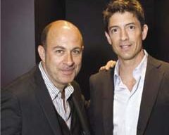 Two men in suits stand side by side outside a stylish boutique on Maiden Lane, both looking at the camera and smiling. The man on the left is bald and wears a striped shirt under his suit, while the man on the right has short, dark hair and sports a white John Varvatos shirt under his suit.
