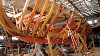 A wooden boat under construction in a workshop embodies craft and tradition. The unfinished hull, supported by metal and wooden braces, reveals the boat's internal frame. The spacious workshop, with scattered tools and materials, boasts a high ceiling featuring exposed beams.