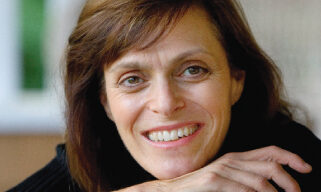 A woman with straight brown hair and light skin, reminiscent of Judy Shils, is smiling and resting her chin on her hands. She is wearing a dark-colored top. The background is blurred and suggests an outdoor setting.