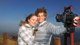 A couple stands in front of the Golden Gate Bridge, smiling for a selfie. The man, holding the camera, wears a striped hoodie while the woman, next to him, wears a white plaid jacket. The bridge and blue sky are clear in the background. Both appear happy and relaxed—Wish You Were Here!