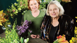 Two women are standing in front of a wooden table covered with a variety of colorful flowers, including lilies, orchids, and tulips. The woman on the left has light red hair and is wearing a green top, while the woman on the right has gray hair and is wearing a dark jacket. Both are smiling, showcasing their Bouquets to Art.