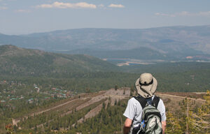A person wearing a backpack and a wide-brimmed hat stands on a hillside overlooking Tahoe’s vast landscape of rolling hills and distant mountains under a clear blue sky with a few clouds. The foreground features green shrubs and scattered patches of snow, embodying the spirit of an outdoor adventure.
