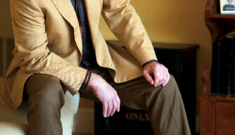 Stephen Denison is sitting on the armrest of a couch, leaning forward with one hand on his knee. He is wearing a beige blazer, brown pants, and a striped shirt. A light brown dog is lying on the floor beside him. The room has a warm, cozy ambiance.