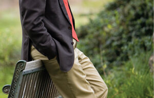 A man in a brown blazer, light blue dress shirt, red tie, and khaki pants leans against the back of a park bench with one foot resting on the seat. He stands on a grassy area in Fairfax with trees and a blurred background. Karl Hoagland captures this serene moment perfectly.