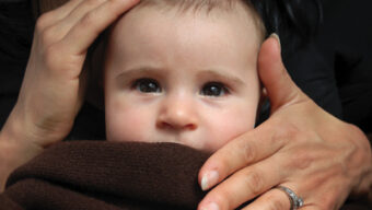 A close-up of a baby looking slightly upwards with wide eyes. The baby's head is gently held by a mother's hand from the top, and another adult's hand is covering the baby's mouth with a blanket. The person has a ring on the finger touching the baby's chin, and hands tenderly cradle the infant.
