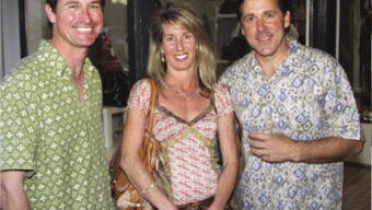 Three people are standing outside a building, smiling at the camera. The person on the left, wearing a green patterned shirt, is holding up five fingers. The person in the middle is wearing a pink and white patterned top, and the person on the right is sporting a blue patterned shirt.