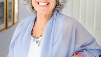A smiling older woman with curly gray hair stands indoors, wearing a light blue shawl over a white top. She has a pink wristwatch and delicate jewelry. Framed pictures hang on a wall behind her, and she appears to be standing next to a staircase railing or shelf, reminiscent of Nancy's List.