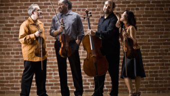 A string quartet stands against a brick wall, smiling and interacting with each other. The group, reminiscent of the Marin Symphony's elegance, includes two violinists, a violist, and a cellist. They hold their instruments and appear in good spirits, suggesting camaraderie and a positive mood.