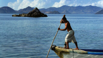 A man stands on the edge of a small boat, using a long pole to navigate through clear, calm waters. He is shirtless and wearing light-colored shorts. In the background, there are mountainous isles under a blue sky with scattered clouds.